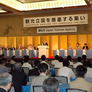 観光庁の英文名を背にあいさつする冬柴国交省。 国会議員、16団体のトップが壇上に並んだ